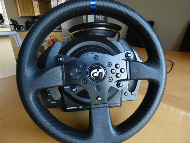 Thrustmaster T300RS GT Edition sim racing wheel on a wooden table, showing the wheel rim face with all the buttons. 