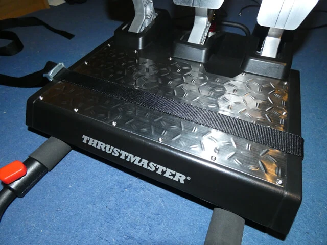 Thrustmaster T-LCM sim racing pedals attached to the Playseat Challenge using a ratchet strap, sitting on a blue carpet.